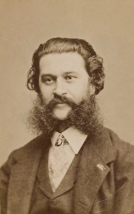 Sepia-toned portrait of a man with curly hair and a thick beard, wearing a dark suit and high-collared shirt.