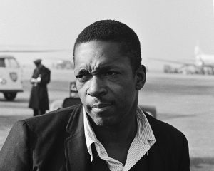 A black and white portrait of a man at an airport, wearing a suit jacket and open-collared shirt, with a serious expression.