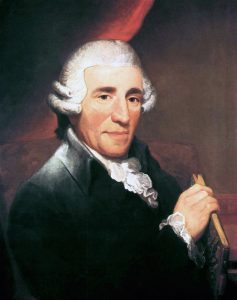 Portrait of an older man with white curly hair in a dark coat holding a book against a dark background.