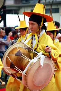 Person in traditional yellow Korean attire playing a janggu drum in a parade.