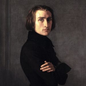 Portrait of a young man with shoulder-length brown hair, wearing a dark coat against a gray background, with arms crossed.