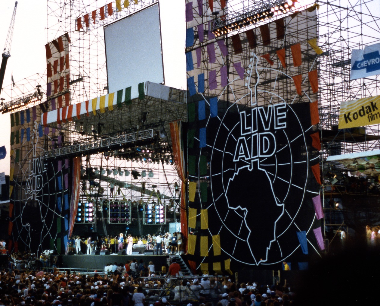 Outdoor concert stage with "LIVE AID" and Africa outline, colorful banners, and a large crowd.