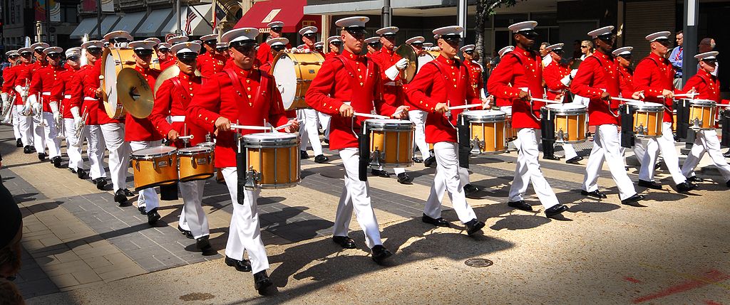 A marching band in red uniforms with white caps playing drums as they parade down a street.