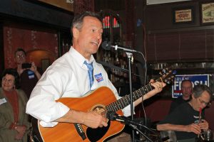A man in a white shirt and blue tie plays guitar and sings into a microphone at a cozy indoor venue.