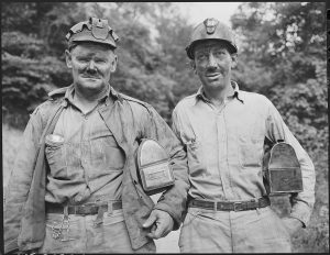 Two miners in dirty work clothes and helmets, holding lunchboxes, standing outdoors.