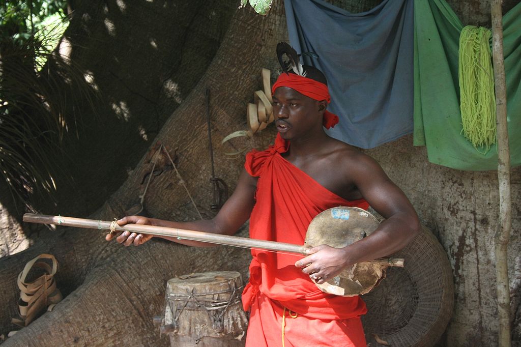 A man in red clothing holds a traditional stringed instrument in front of a large tree trunk with hanging cloths and drums nearby.