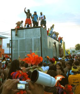 A vibrant street scene during a festival, featuring a crowd in colorful costumes and performers atop a large vehicle.
