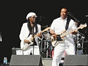 Two musicians in white outfits perform on stage with electric and bass guitars.