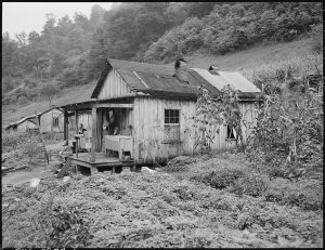 Rustic wooden house in a rural area with overgrown plants and a small porch where two people sit.