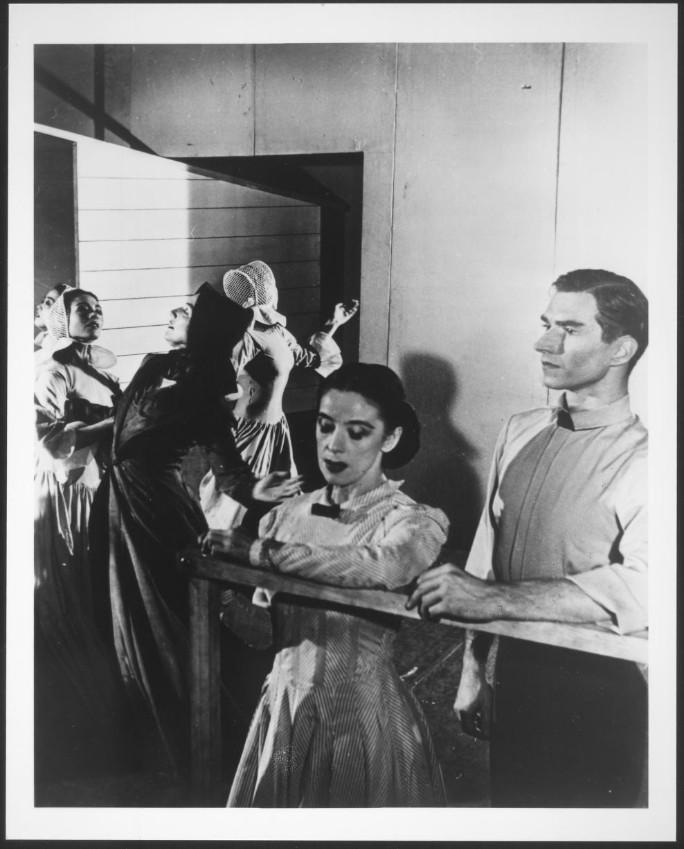 A group of dancers holding a barre in a studio setting, dressed in period costumes.