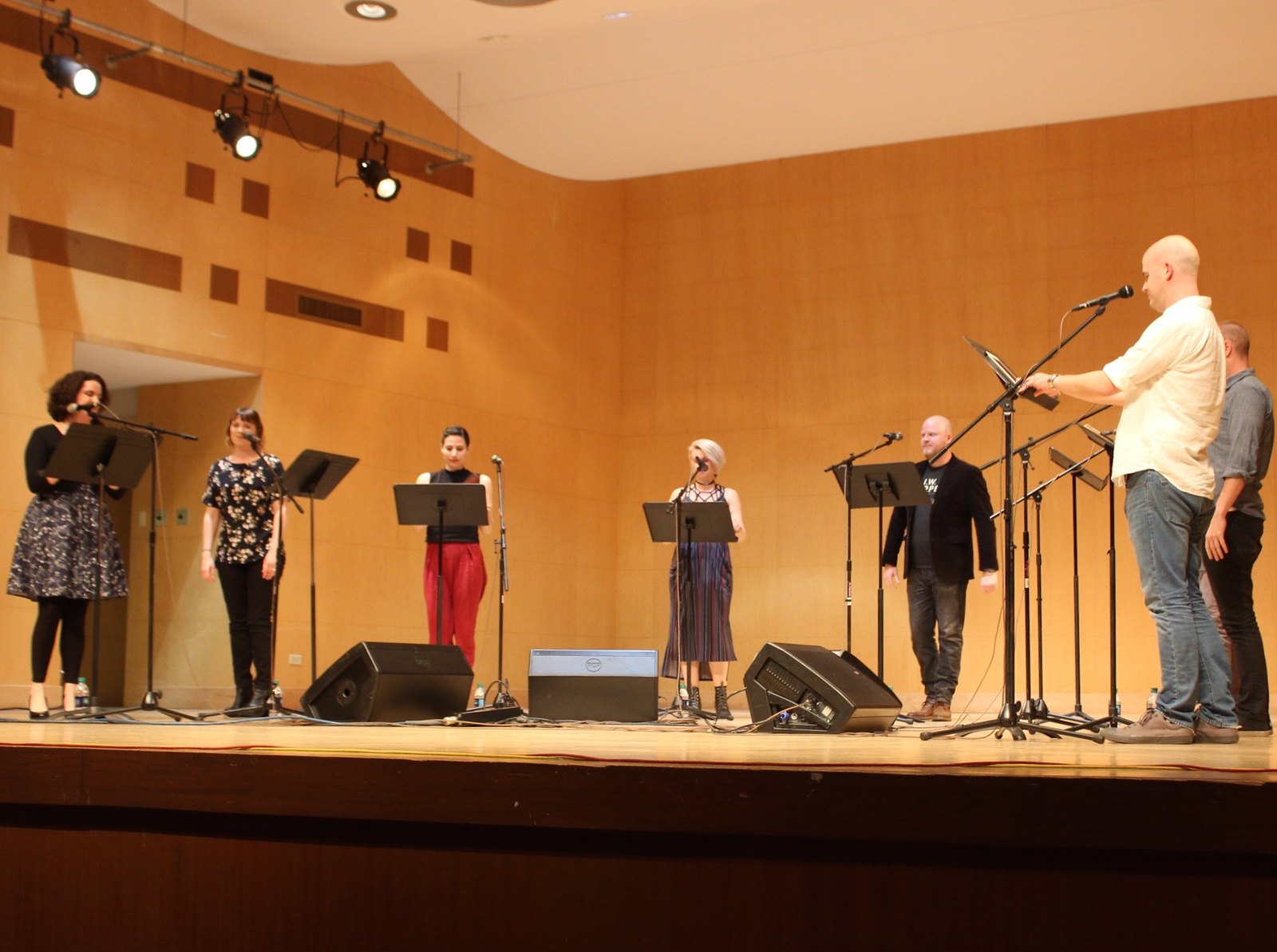 Seven people stand on a stage with microphones and music stands in a concert hall.