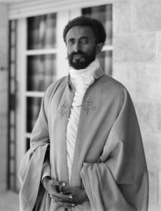 A man in an embroidered robe stands indoors near a window.