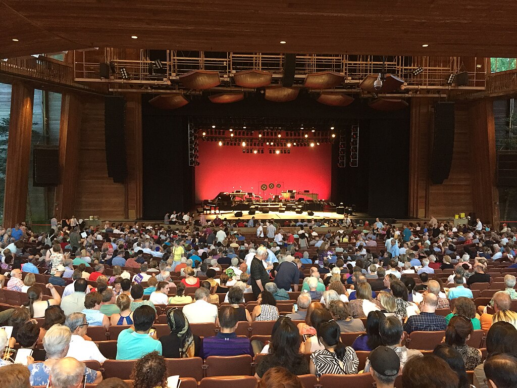 A packed indoor theater with a wooden interior, featuring a red-curtained stage set with musical instruments.