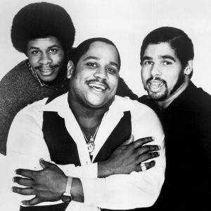 Three men smiling, with one in the foreground wearing a white shirt and dark vest, flanked by two others in casual attire.
