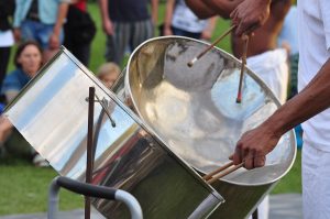 Person playing a steelpan drum with drumsticks in an outdoor setting.