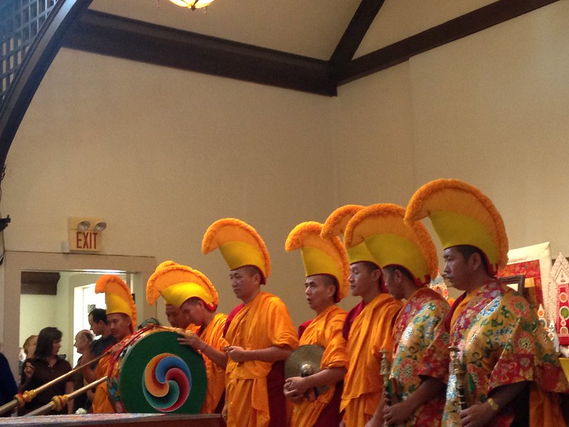 A group of Tibetan Buddhist monks wearing orange robes and yellow hats, holding musical instruments inside a room.