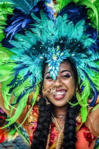 A person with a vibrant blue and green feathered headdress, smiling with gold facial jewelry and colorful makeup.