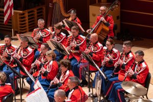 Military band in red uniforms performing on stage with woodwind and brass instruments.