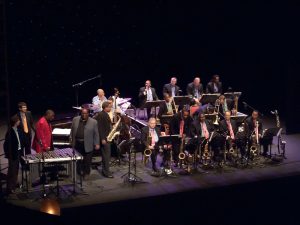 A jazz band performing onstage with various musicians playing wind and percussion instruments against a dark backdrop with star-like lights.