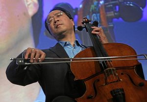 A musician playing a cello on stage with a large projected image of his face in the background.