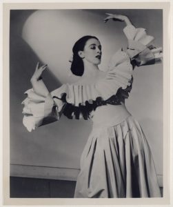 A black and white photograph of a dancer in a dress with ruffled sleeves and pleated skirt, posing gracefully with raised arms.